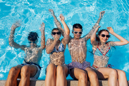 Cute teens in swimsuits and sunglasses in the pool. Smiling boys and girls dive into the clear water. Joyful teenagers relax and have fun together.の写真素材