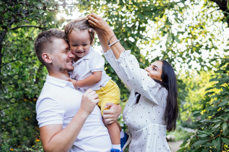 Cute family in light casual clothes in garden on terrace of house. Young couple and their outdoor sons on summer sunny day. Dad standing back and holding his toddler boy. Attractive woman is smiling.の写真素材