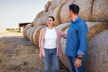 A young married couple of farmers in casual clothes stand near rows of haystacks outdoors in the country. Husband and wife work and live on a farm. Harvestingの写真素材