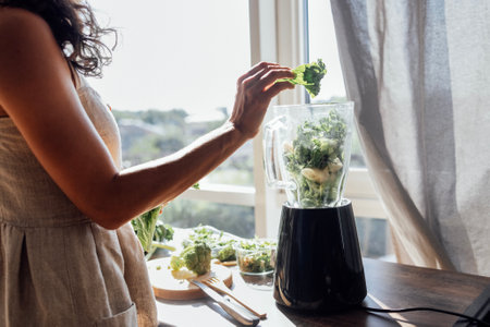 A close-up of a blender with green vegetables and herbs and a female hands with a leaf of wavy cabbage. The girl makes a vitamin smoothie at home. Broccoli and arugula on the kitchen table.の写真素材