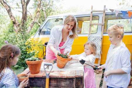Young woman and her three children are planting flowers in pots in backyard. Pretty mother, cute girls and boys are engaged in gardening. Happy family has time together. Yellow minivan on background.の写真素材