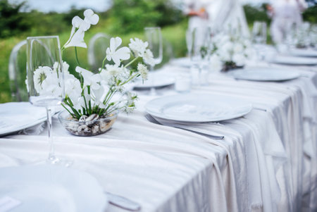 The festive table with a white tablecloth is decorated with flowers. Empty plates and glasses waiting for the guests. A holiday lunch in the garden. An open-air restaurant or cafeの写真素材