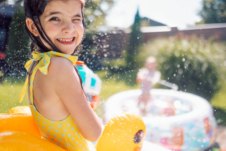 Joyful little girl in swimsuit with inflatable circle on terrace. Cute kids are messing around and splashing water in the backyard of the house. Carefree children have fun in pool in summer outdoorsの写真素材