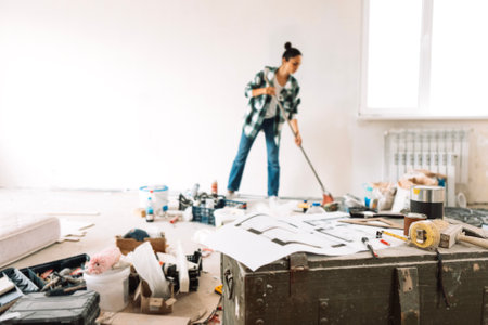 Renovation of the apartment. A young woman with a sweeping brush cleans the room from construction debris. A girl in casual clothes sweeps the floor with a broom. Construction materials and tools.の写真素材