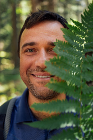 Close-up portrait of young happy man with fern in nature. Smiling tourist hides his face under green...