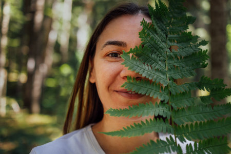 Close-up portrait of a young woman with a fern in nature. A smiling girl hides her face under a...