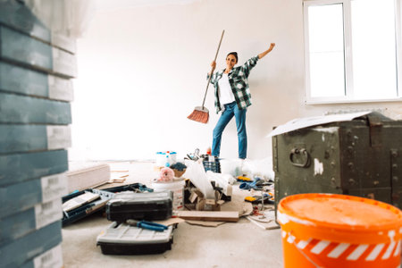 Young smiling woman with sweeping brush is dancing in renovated room. Break between cleaning of construction debris. Carefree girl in casual clothes with broom. Construction materials and tools.の写真素材