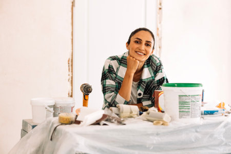 The laughing woman at the table with construction tools. A smiling girl in casual clothes is making repairs in the apartment. A worker among building equipment and materials. Home renovationの写真素材