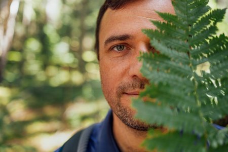 Close-up portrait of young happy man with fern in nature. Smiling tourist hides his face under green leaf in forest. Carefree parson in casual clothes outdoors in woods. Travel and active recreationの写真素材