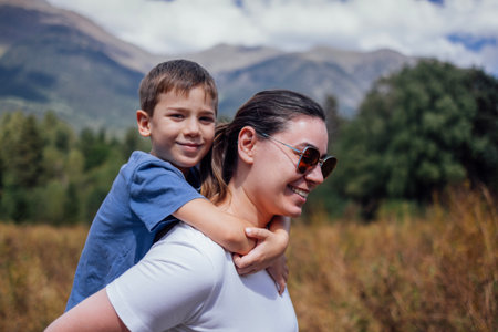 A young woman carries her little son on her back in nature. A mom in a white T-shirt and sunglasses enjoys the views of the mountains with her kid. A family of tourists on a walk through the highlandsの写真素材
