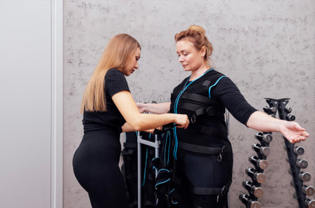 Female coach puts on suit for ems workout on senior woman. The trainer fixes the wires on clients chest. Preparing for electrical muscle stimulation training in fitness studio or gym. Copy space.の写真素材
