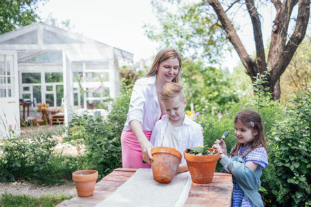 Young woman and her two children are planting flowers in clay pots in the garden. An attractive mother, a cute girl and a boy in the backyard. A happy family has a good time together.の写真素材