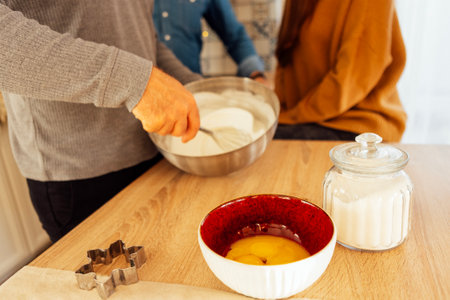 Close-up of a male hands moving a whiskey of white cream in a metal bowl. Plates of eggs, a jar of sugar and a cookie mold are on wooden table. Making Christmas cookies.の写真素材
