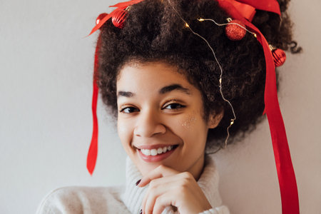 Close-up of young African with curly hairstyle decorated with red ribbon, garland and Christmas balls. Cute black girl in warm sweater is getting ready for New Year party. Woman enjoying holidayの写真素材