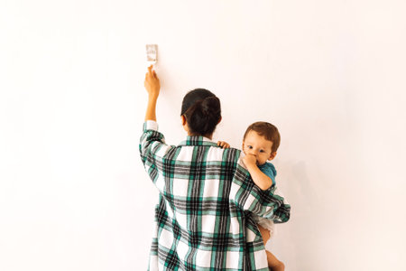 A young woman holds a child and paints a wall. The view from the back. A mother with a small kid is making repairs in an apartment. Mom and baby apply paint to the wall of the house with a brush.の写真素材