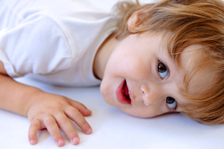 A close-up portrait of a small carefree child lying on the floor. A cute smiling kid in casual clothes is playing at home. The happy sweet boy laughs and looks at the camera. White backgroundの写真素材