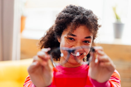 A young African woman smiles and keeps her glasses at home. A short-sighted attractive black girl laughs and wipes her glasses indoors. A cute female teenager checks the cleanliness of the lensesの写真素材