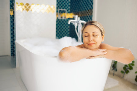 A young woman has closed her eyes and is taking a bubble bath. The blonde relaxes in hot water. A charming girl is smiling and relaxing at home. Stylish modern bathroom interiorの写真素材