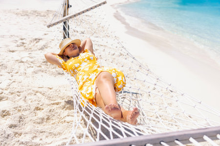 A young woman in a straw hat and a summer dress is lying in a hammock near the ocean. The lady sleeps and relaxes on the seashore. The girl is enjoying her vacation and the sea breeze on the beachの写真素材