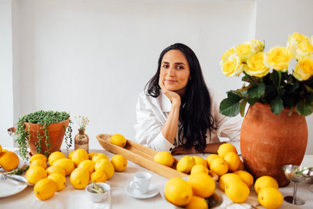 Young woman smiles and sits at set table with lemons. Clay pitcher with yellow roses, houseplant, kitchen utensils on tablecloth. Calm brunette girl in rustic vintage interior. Fragrant citrus fruitsの写真素材