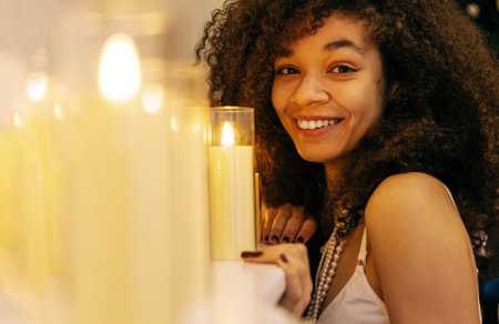 A close-up of a young smiling African woman at home. A charming laughing black girl near lighted candles. An attractive teenager in an elegant festive dress and a pearl necklace in the living roomの写真素材