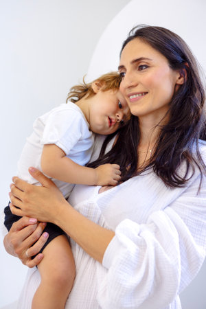 Close-up of a cute baby with a charming mom in casual clothes. A young attractive woman smiles and hugs her little son. The brunette holds a surprised sweet child. White isolated backgroundの写真素材
