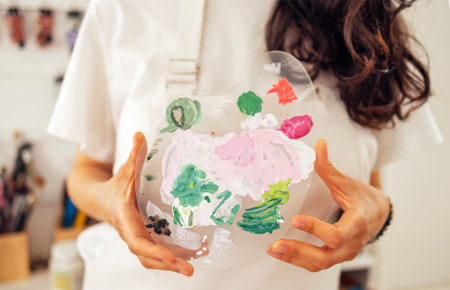 Close-up of well-groomed female hands holding plastic palette with mixed colors. Young woman in casual clothes and apron in art studio. Girl at painting workshop. Creative activity or hobbyの写真素材