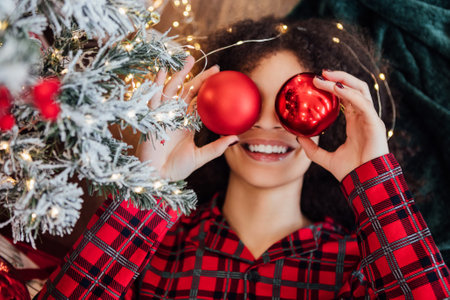 Close-up funny portrait of young happy African woman in plaid pajamas with red Christmas balls. Cute black girl is laughing and decorating Christmas tree at home. Preparing for holiday. Cozy interiorの写真素材
