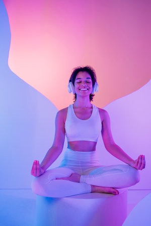 A young African in white sportswear sits in a meditation pose on round platform. A smiling black girl enjoys music with headphones. Relaxed and happy woman on podium. Vibrant neon colorful backgroundの写真素材