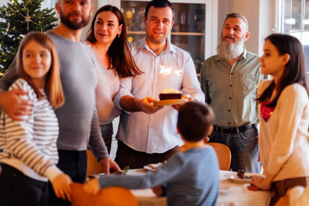 A big happy family at a festive table at home. An attractive young man with a cake with lighted...