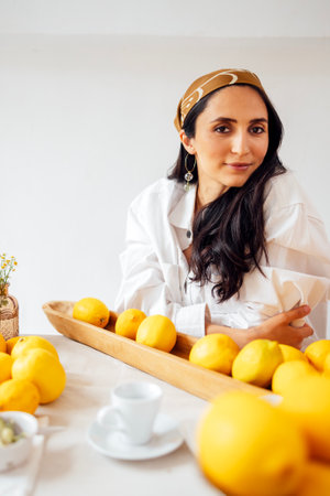 Young woman with headscarf is smiling and sitting at set table with lemons. Kitchen utensils and daisies on tablecloth. Calm brunette in shirt in vintage rustic interior. Fragrant citrus fruitsの写真素材