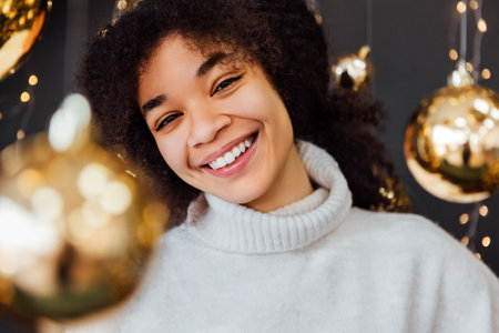 Young African woman with curly hair smiling and standing among golden Christmas decorations. Laughing black girl in cozy white sweater. Lady is enjoying the festive atmosphere. Shiny balls and garlandsの写真素材