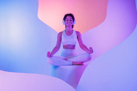 A young African in white sportswear sits in a meditation pose on round platform. A smiling black girl enjoys music with headphones. Relaxed and happy woman on podium. Vibrant neon colorful backgroundの写真素材