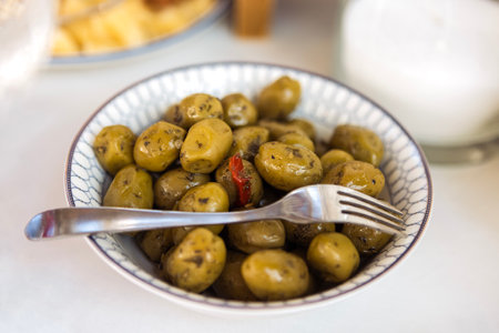 Green olives with spices. A close-up of a plate with delicious fruits of the olive tree and herbs on a white table. Tasty vegan snacksの写真素材