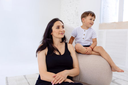 A young pregnant woman in a black dress is sitting with her son on the couch. Adorable mom and her little boy on the sofa. A beautiful lady waiting for a baby with her cute child at homeの写真素材