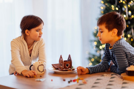 Small children decorate the gingerbread house in the dining room. Cute brother and sister drink milk and eat traditional Christmas cookies at home. Cozy interior with a Christmas tree.の写真素材
