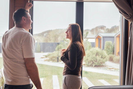 An attractive man and woman stand by the window in the living room. A happy married couple enjoys a conversation in their modern home. The charming wife smiles as she chats with her husbandの写真素材
