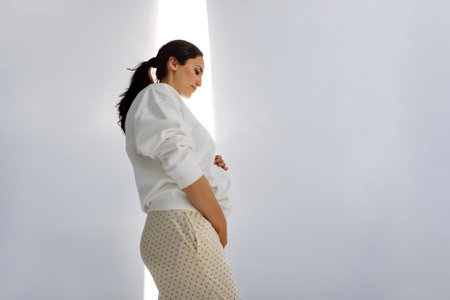 A pregnant woman gently holds onto her belly. The expectant mother touches her tummy against the background of a white wall. Bright sunlight. An attractive lady waiting for a baby indoorsの写真素材