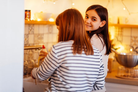 Two cute teenage girls are washing dishes after cooking at home in the kitchen. Smiling sisters are doing their homework. Friends spending time and working together. Cozy interior with Christmas decorの写真素材