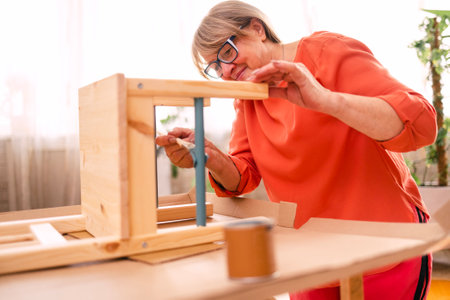 An elderly woman in orange casual clothes is painting a wooden chair in the living room. A mature...