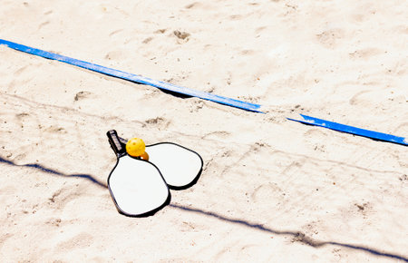 Two pickleball paddles and a yellow ball rest on a sand outdoor court. An energetic sports game on a clean beach. Copy spaceの写真素材