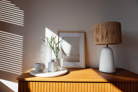 A wooden chest of drawers and a window with louvered shutters. An elegant table lamp, poster frame mock-up, a tray with a glass jug and a vase, scented candles and cup. Cozy details of the interiorの写真素材