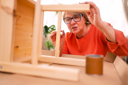 An elderly woman in orange casual clothes is painting a wooden chair in the living room. A mature woman with glasses makes furniture. A satisfied pensioner is engaged in her favorite hobbyの写真素材
