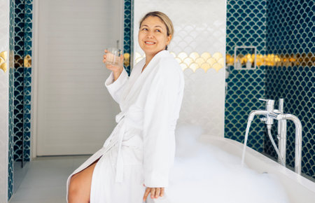 Young woman in white bathrobe holds glass of water and takes bubble bath. Charming blonde is smiling...