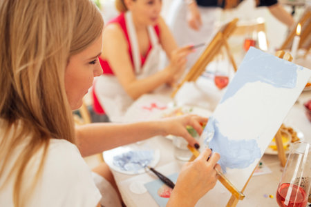 Close-up of canvas on easel and hand with spatula. Young artist paints picture at table. Blonde in painting workshop. Girl is learning to draw in art studio. Creative activity and hobbyの写真素材