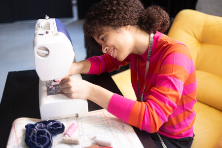 Young African woman working at sewing machine. Charming black girl in red sweater with measuring tape. Materials for sewing and fabric patterns in workplace. Seamstress is engaged in needleworkの写真素材