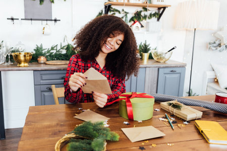 Cute African woman in pajamas is smiling and packing greeting cards into envelopes. Charming black girl is wrapping New Year gifts at table in kitchen. Cozy home interior with Christmas decorationsの写真素材