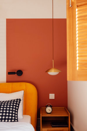Close-up of wooden laconic classic bedside table with book and alarm clock. Cozy interior with bright accents. Bed with contrasting pillows. Stylish wall lights. Open window with louvered shuttersの写真素材