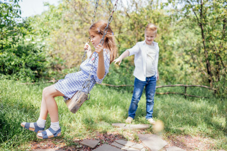 Pretty schoolboy in jeans and white shirt is rocking his younger sister on swing.