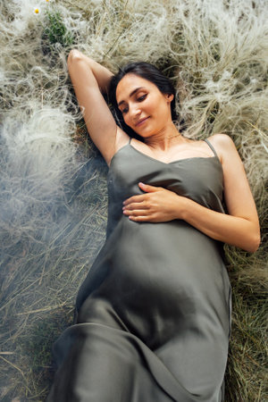 Young pregnant woman in green satin dress lies on grass in summer. Charming expectant mother gently holds her belly in meadow. Peaceful woman waiting for a baby among flowers relaxes in clearingの写真素材